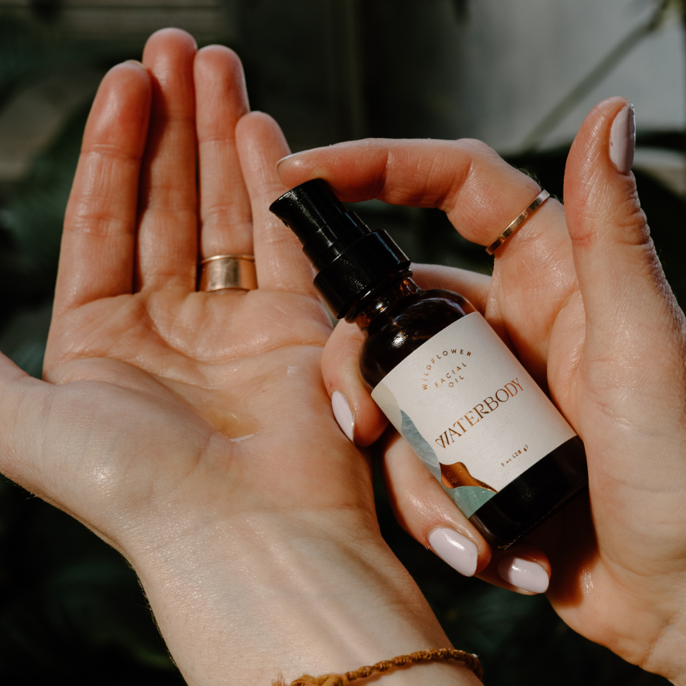 A woman wearing gold rings holds a bottle of facial oil in one hand and pumps a few drops of the oil into her palm