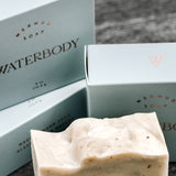A stack of soap boxes are blue and rectangle in shape. A bar of soap sits at the foreground