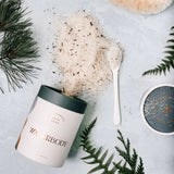 A tube of bath salts spills out, displaying salts flecked with forest plants and botanicals. The image is framed with evergreen boughs, forest ferns, and a small white spoon