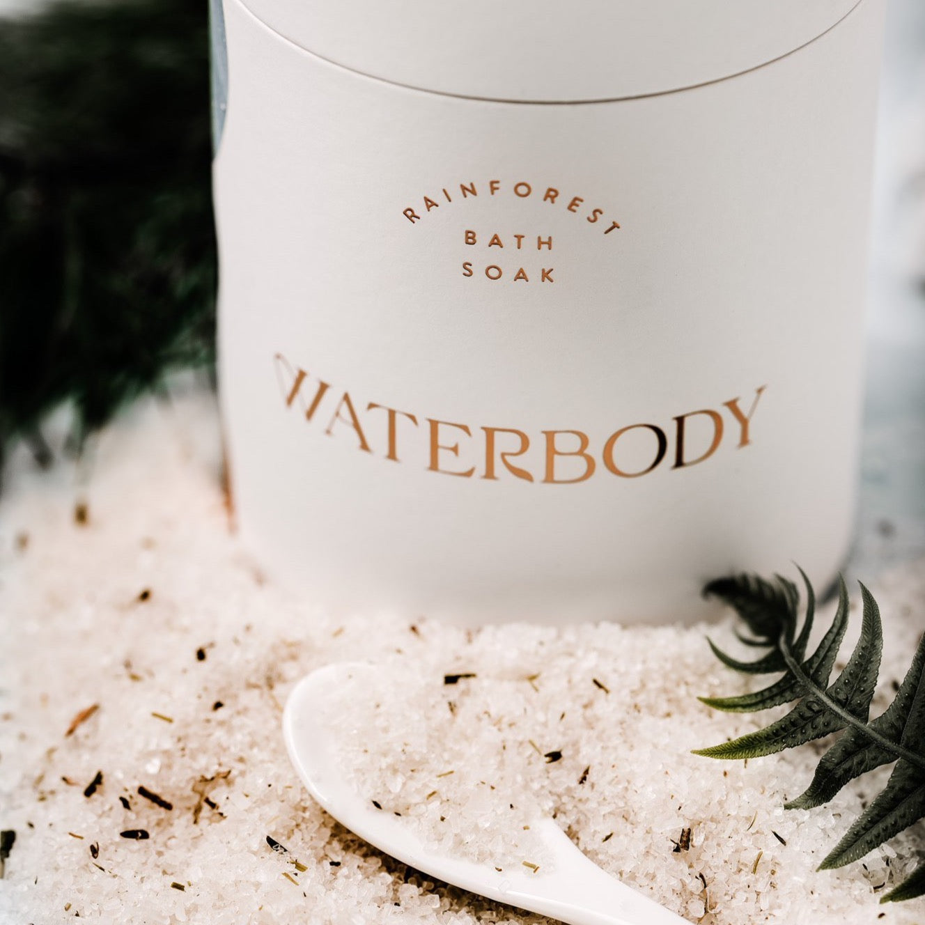 A tube of bath soak is framed by a spilled pile of the bath salts, flecked with botanical forest plants. The image is framed with a white spoon and green fern frond