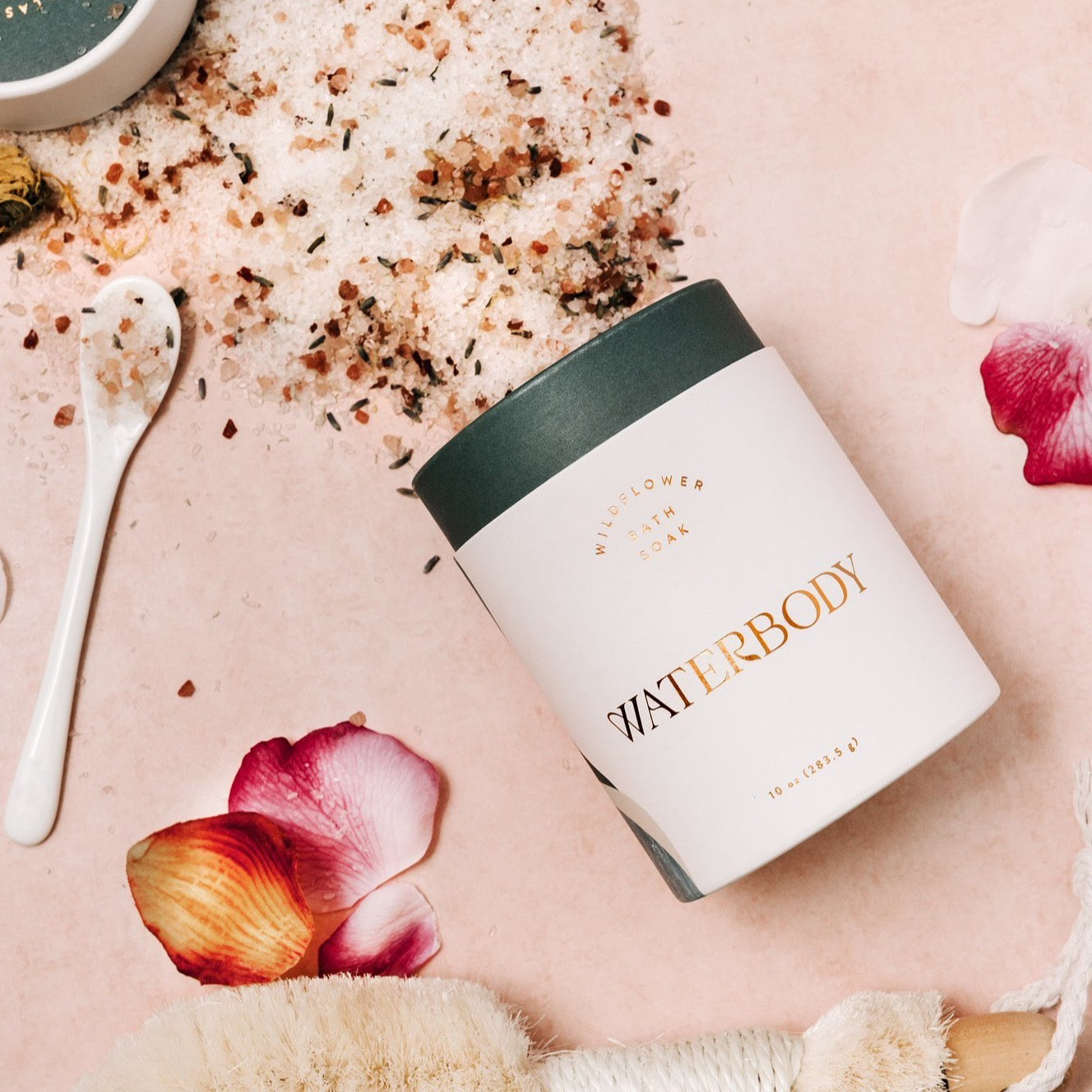 A white and jade green tube of bath salts is spilled out over a faint pink backdrop. The salts are flecked with pink salt and botanical flower petals. The image is framed with rose petals, a small white spoon, and a dry body brush