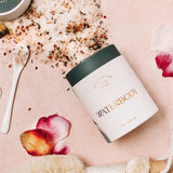 A white and jade green tube of bath salts is spilled out over a faint pink backdrop. The salts are flecked with pink salt and botanical flower petals. The image is framed with rose petals, a small white spoon, and a dry body brush
