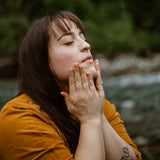 A woman applies facial oil to her skin beside a natural stream