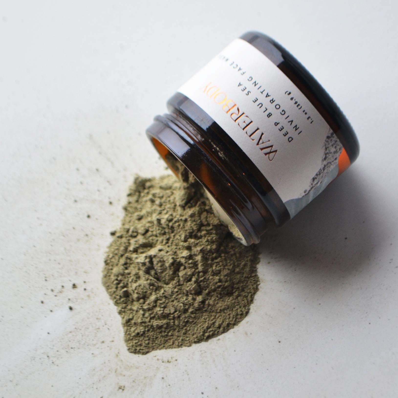 A brown glass jar of Waterbody Deep Blue Sea Face Mask is open and spills out over a white surface. The mask is a dry sea green powder
