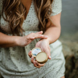 A woman with long brown hair and a green polka-dot dress sits beside the ocean and applies an open tin of Calm Body Balm to the inside of her wrist