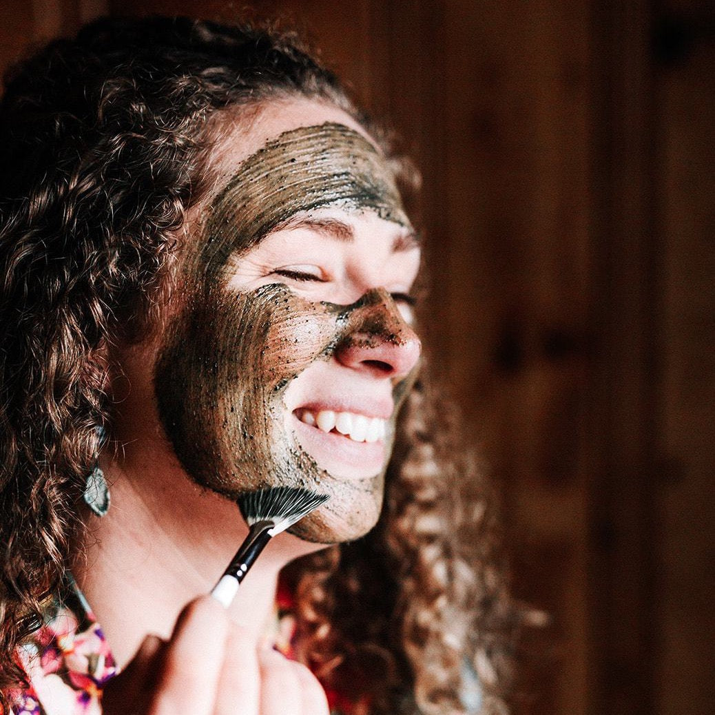 A woman with brown curly hair smiles as she applies a blue-green face mask treatment to her sin with a small brush