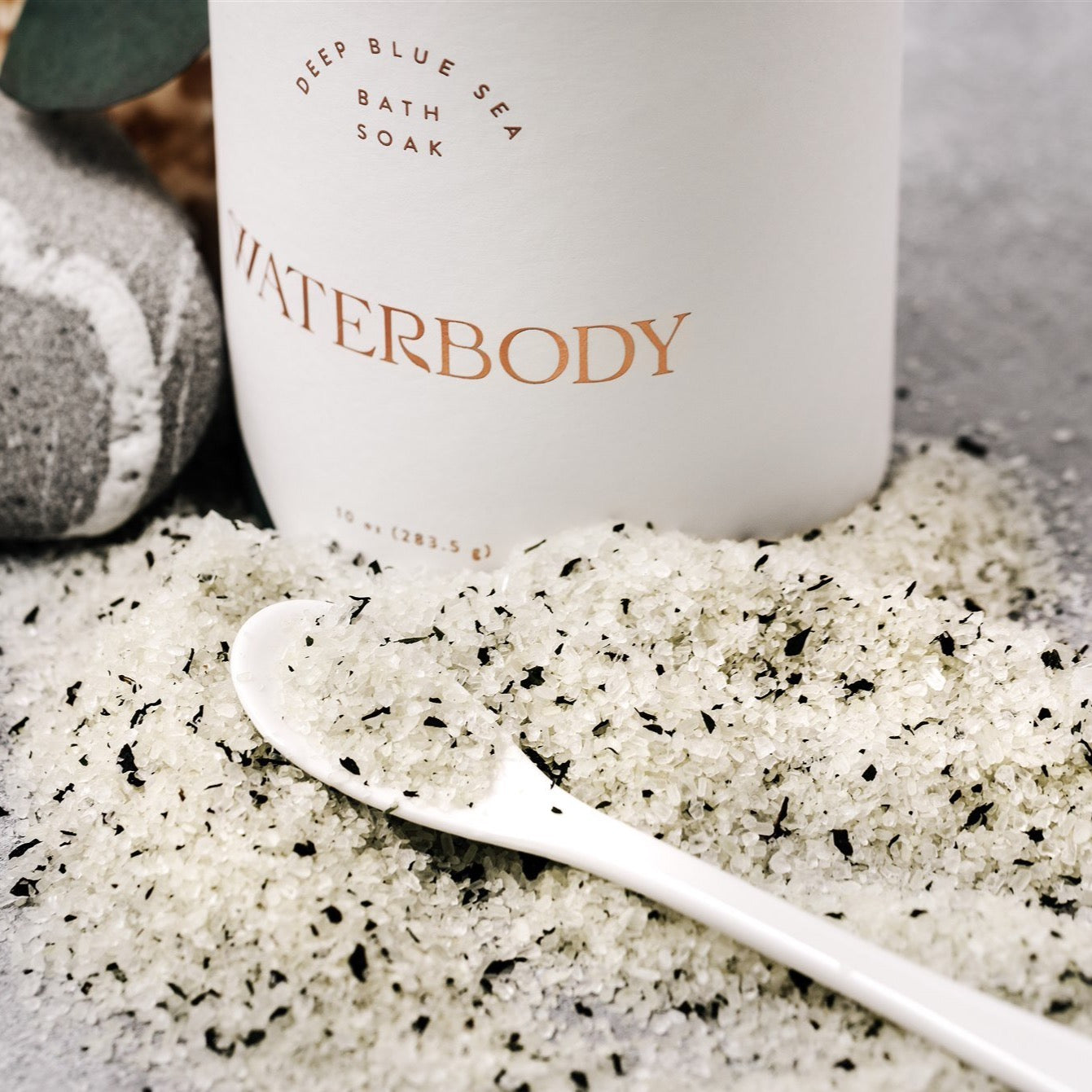 A white paperboard tube of Deep Blue Sea Bath Soak packaging with gold foil lettering sits beside a mound of the bath salts peppered with flakes of kelp and peppermint.