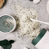 A paperboard tube of Deep Blue Sea Bath Soak is spilled out over a grey stone backdrop. The bath salts are peppered with flakes of plant material, and a white spoon, stone, and sea sponge are staged to frame the photo