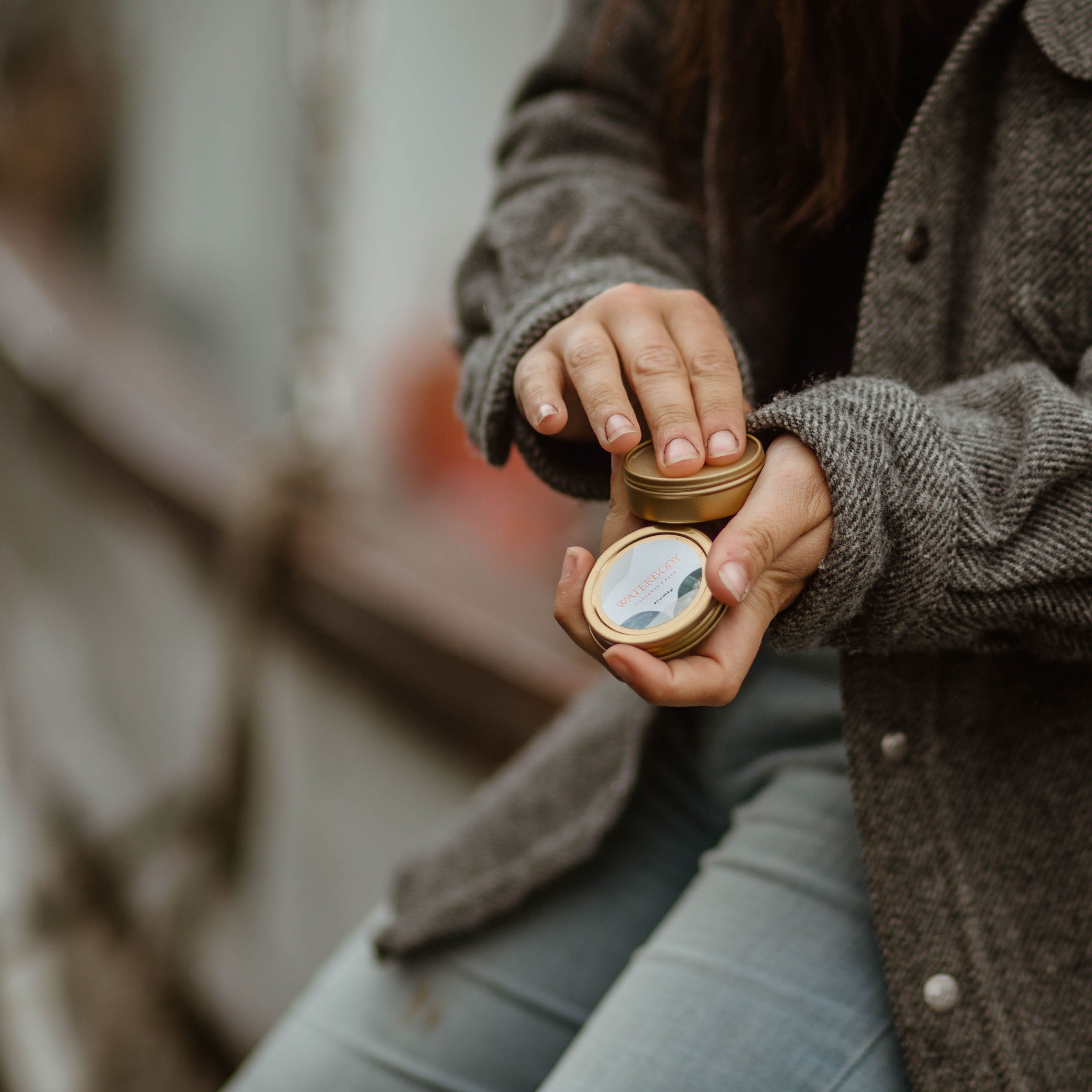 A woman wears a wool fisherman's coat and jeans sitting beside a commercial fishing boat in Alaska. She holds a round gold tin of hand salve and dips her fingers in to apply the product