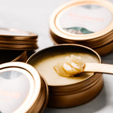 A round golden tin of salve is open with the lid to the side. A small golden spatula is dipped into the product to show a creamy golden butter like texture. 