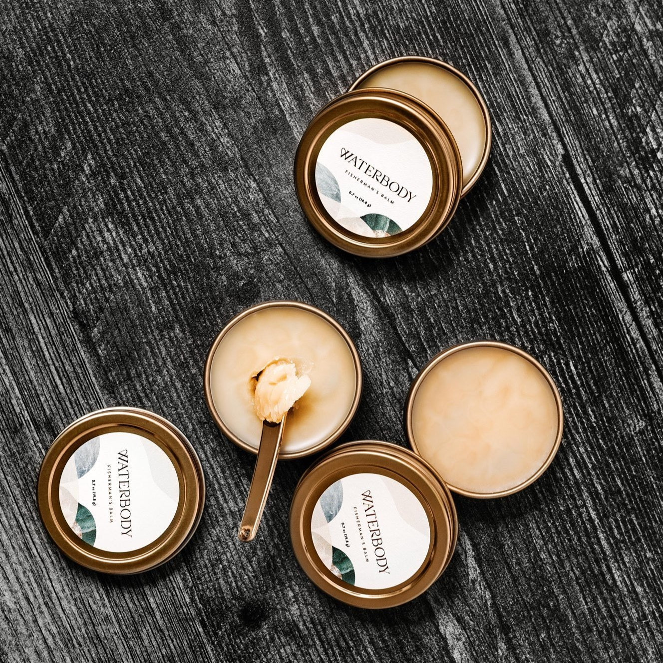 Three tins of hand salve sit opened against a grey wood backdrop. The tins are round, golden, and hold a buttery textured balm.