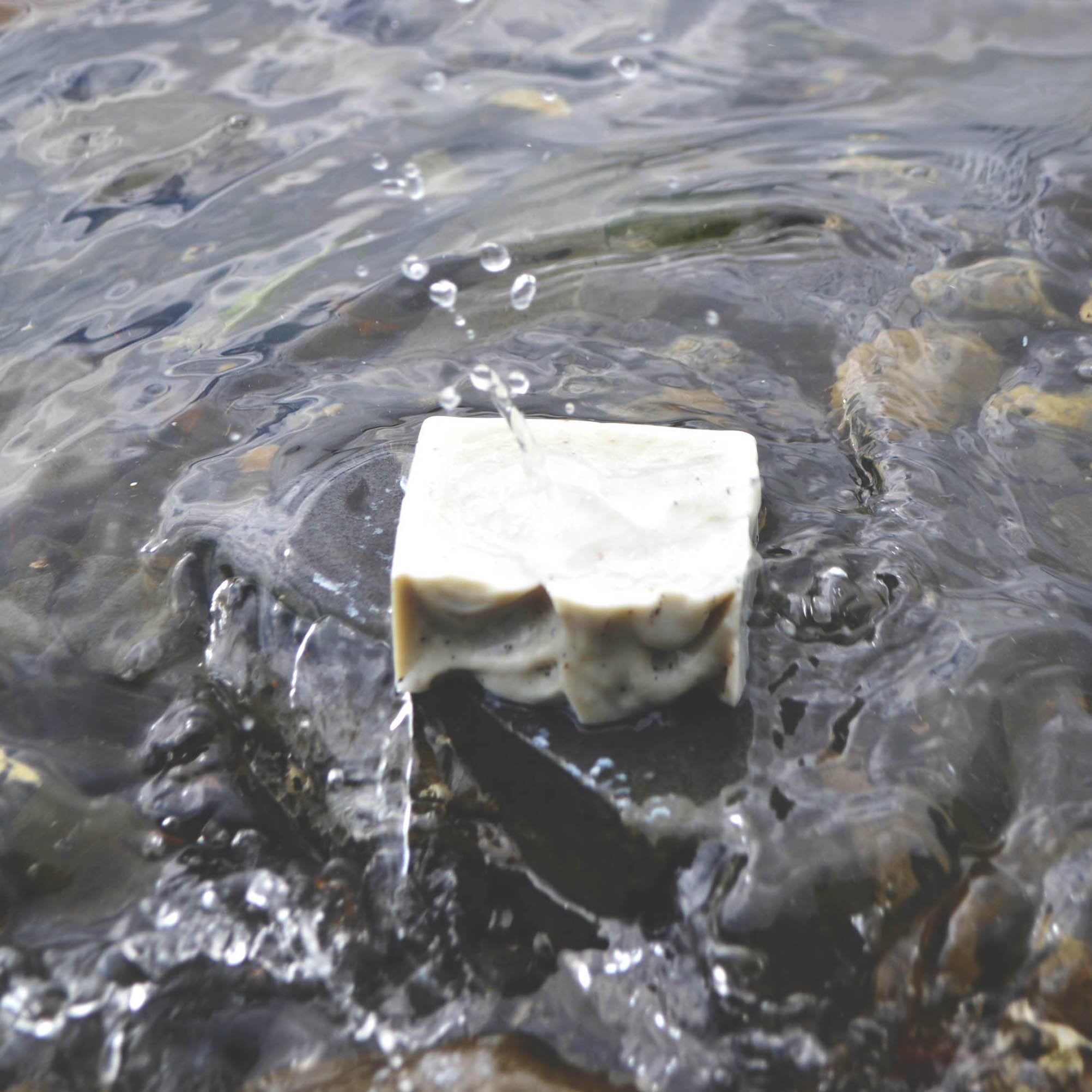 A blue-grey bar of soap sits on a rock at the shore of the ocean. It is lightly splashed by a wave.