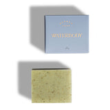 A rectangle blue box sits beside a rectangle bar of greenish colored soap flecked with bits of kelp. the background is white