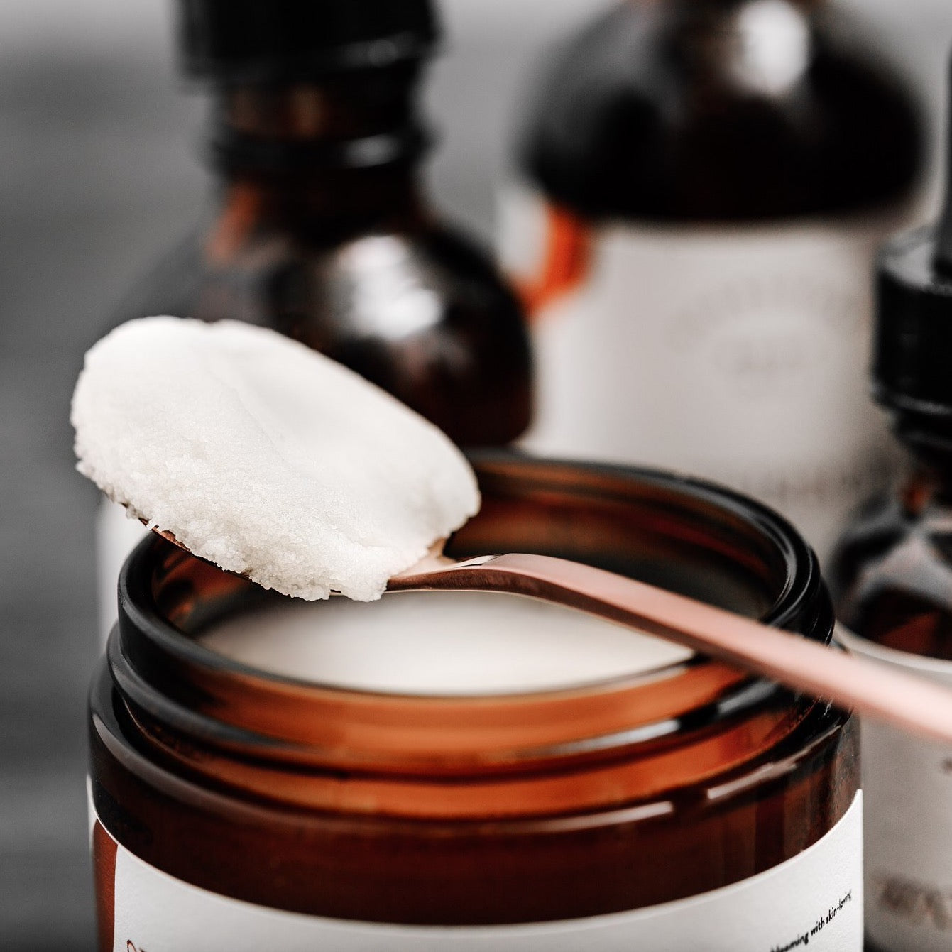 A copper spoon filled with a scoop of cream colored body butter sits atop a brown glass jar of product. Blurred out bottles of skincare products are in the background