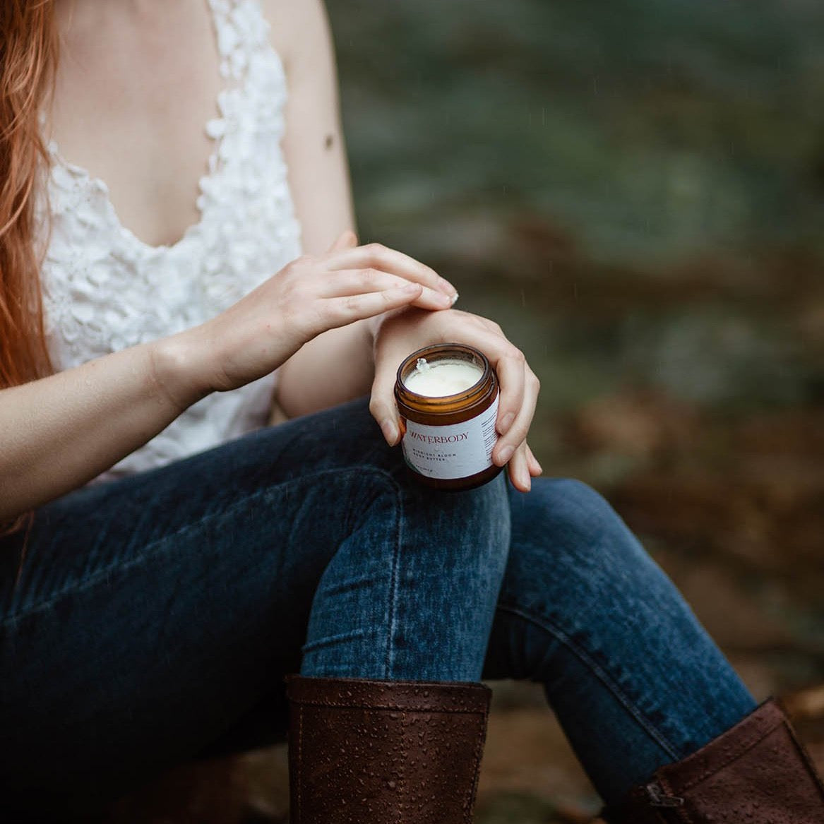A woman sits beside a stream in the forest. She is wearing a white lace tank top, blue jeans, and brown boots. She holds a jar of body butter in her hands and lightly applies the product to her skin.