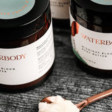 Jars of body butter sit in the background, while a copper spoon holds a scoop of cream colored body butter