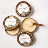 Three tins of Waterbody Calm Body Balm sit against a white marble backdrop. One tin is opened with a small gold spatula in the product to show a thick buttery salve-like texture. The tins are gold with white, blue, and green packaging.