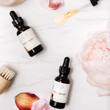 Two bottles of facial oil lay against a white marble backdrop. There is a glass dropper of oil spilling its golden liquid contents, as well as a rose, rose petals, and small facial brush in the image