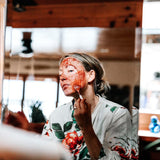 A woman applies a magenta colored face mask treatment to her skin using a small white brush. She looks into a mirror wearing a yellow flowered bathrobe.