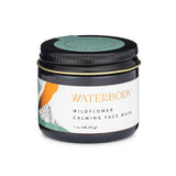 A brown glass jar with a white label that reads "Waterbody wildflower calming face mask" in gold text has a black metal lid and jade green seal. It sits against a white backdrop
