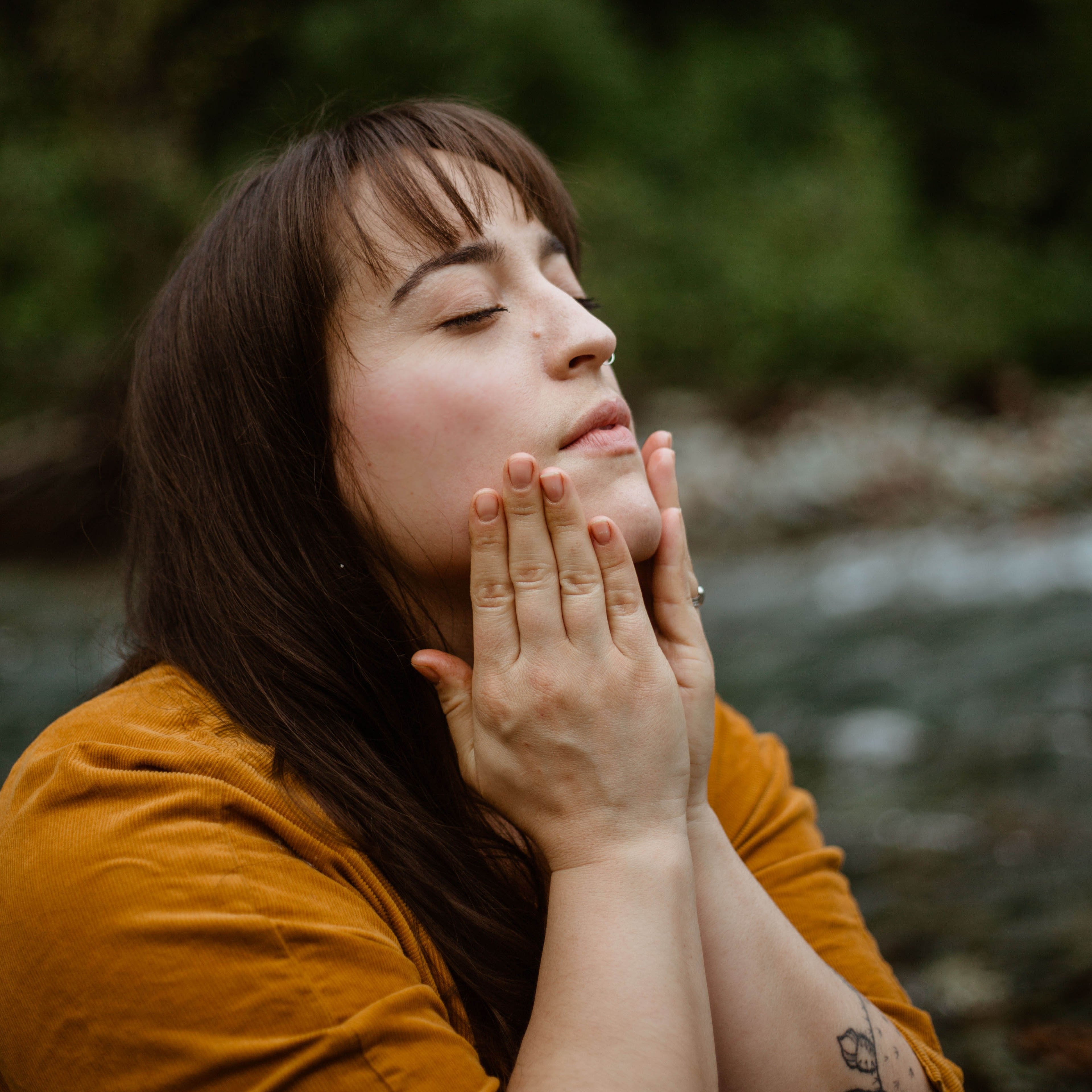 A woman with long brown hair wearing a mustard yellow shirt is outside near a forested stream. Her eyes are closed and she is pressing her hands to her face as though applying a skincare product.