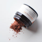 A brown glass jar of face mask spills out onto a white surface. The product is a deep magenta powder