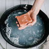 A hand holds a bar of pink clay Alpenglow Soap over a black ceramic bowl of water. A sprinkle of water pours over the bar of soap creating suds and lather in the bowl