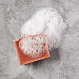 A pink clay bar of Alpenglow soap sits against a grey marbled backdrop with a foaming soap lather